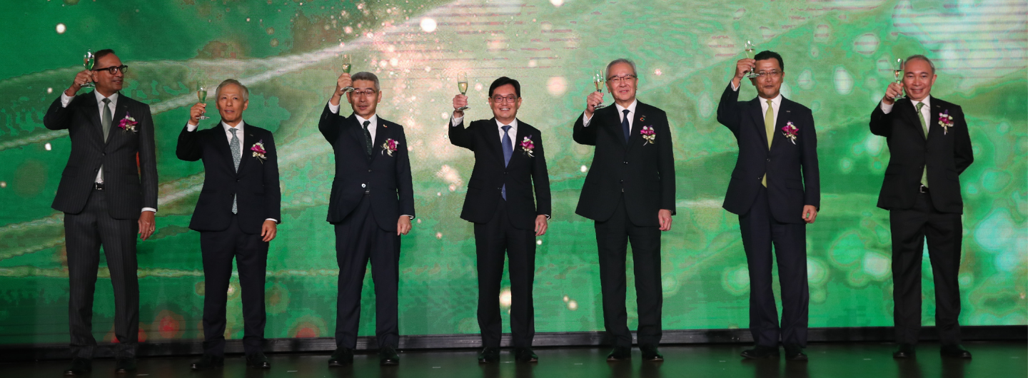 Seven people in suits stand on a stage, each raising a champagne glass. Green patterned backdrop.